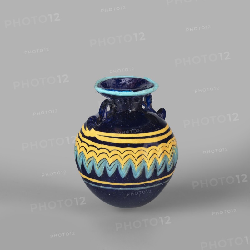 Globular aryballos, Egyptian workshop attributed, globular aryballos decorated at the widest point of the body with a vertically grooved band of zigzag decoration, changing into spirals at the bottom and top of the body. Navy blue glass; thread decoration, embedded blue and yellow; 6th–4th century BC; place of origin, aryballos vessel; weight 0.05 kg; height 6.5 cm; diameter 5.6 cm., A small rounded ceramic vessel with a flared rim and two small loop handles, predominantly deep blue in color with bands of yellow and aqua decorative zigzag and stripe patterns around its midsection; the rim has a lighter turquoise blue edge, and the background is a uniform light gray.