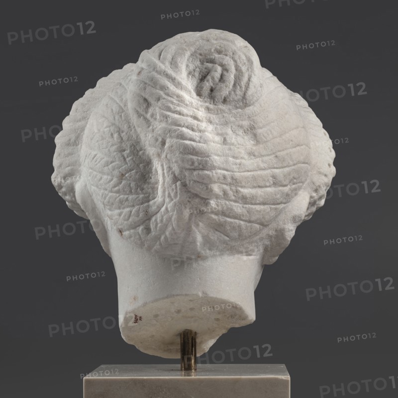 Portrait of a Roman woman, Head with a broad face and an elaborate hairstyle: hair twisted into small curls marked with a gimlet; the rest gathered at the back in a small bun. Round, full face, with a triangular forehead. Roman sculpture, female portrait, masterpiece, ca. 82–92 CE, Rome, Roman Empire 27 BC–476, sculpture. Dimensions: height 30.5 cm, width 27 cm, depth 25 cm; weight 36.5 kg., A light-colored sculpted object viewed from behind showing a textured, rounded top with layered, ribbed patterns and carved folds forming a central spiral; the lower portion tapers into a smoother neck-like base mounted on a vertical metal rod and set into a rectangular block; visible colors include shades of white, off-white, gray, dark gray, and a few small brownish spots.
