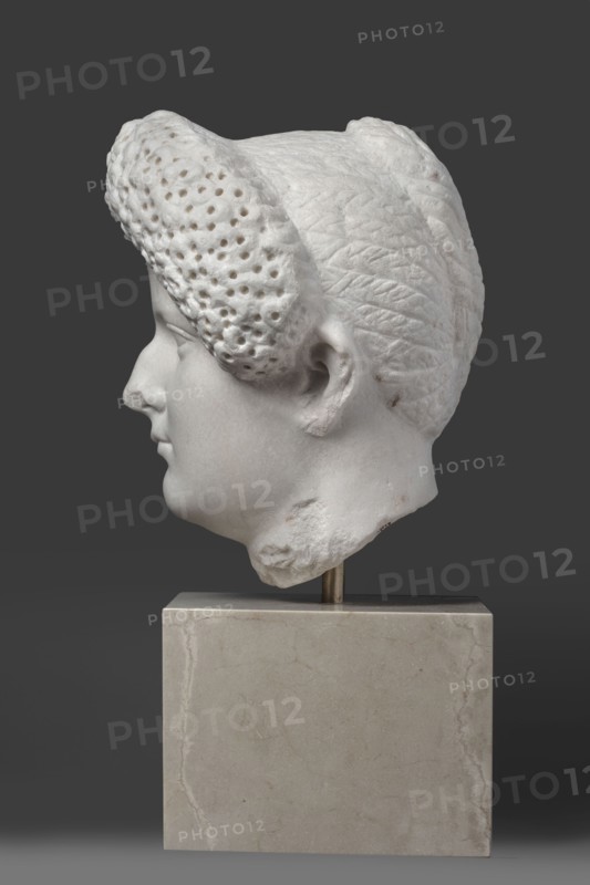 Portrait of a Roman woman, Head with a broad face and an elaborate hairstyle: hair twisted into small curls marked with a gimlet; the rest gathered at the back in a small bun. Round, full face, with a triangular forehead. Female portrait, Roman sculpture, hairstyle, masterpiece. Date: 82–92 CE, place of origin possibly Rome, Roman Empire 27 BC–476. Marble sculpture; height 30.5 cm, width 27 cm, depth 25 cm, weight 36.5 kg., A white marble sculpted head in profile mounted on a rectangular pale beige stone base, shown against a solid dark gray background; the head features textured hair with a perforated curl at the front and a braided or patterned section toward the back, a smooth facial surface with a defined nose, lips, ear, and jawline, and some rougher damaged areas on the lower neck, with small brownish discolorations on parts of the marble.