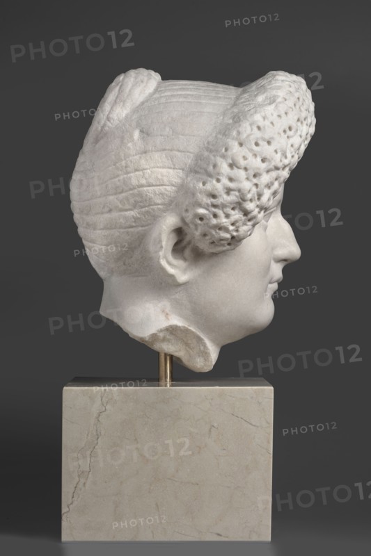 Portrait of a Roman woman, Head with a broad, round full face and a triangular forehead; elaborate hairstyle: hair twisted into small curls marked with a gimlet, the rest gathered at the back in a small bun; female portrait, Roman sculpture, masterpiece; date circa 82–92; origin Rome, — Roman Empire 27 BC–476; sculpture; height 30.5 cm, width 27 cm, depth 25 cm, weight 36.5 kg, A white marble sculpted head shown in profile mounted on a beige marble rectangular base with a short metallic support, the head displaying stylized textured hair with ridged and perforated sections, a visible ear, forehead, nose, lips, and chin, all against a solid dark gray background; colors visible include white, beige, metallic gold, and dark gray.