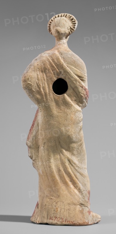 Figurine of a maenad. A young woman standing in a wreath tied with a ribbon is dressed in a chiton slipped from her right shoulder exposing her right breast and upper arm, and a cloak that envelops her hands. The left hand rests on her hip; the right leg is pushed back. The robe is light pink and decorated at the bottom with a wide border and a narrow beetroot-colored stripe above it. A similar border is found at the bottom of the cloak, and a wide border in the same color runs around the cloak. The back of the figurine is unfinished. Without base. 3rd–2nd century BC previously dated 2nd century BC. Myrina. Figurine. Height 26.5 cm; width 9.2 cm; depth 7.4 cm; weight 0.39 kg., A worn statue viewed from the back showing a standing human figure draped in flowing garments with visible folds and a rounded halo-like element around the head; the surface shows a patina of beige and tan with areas of faded reddish pigment and small darker spots, and there is a dark circular hole in the upper b...