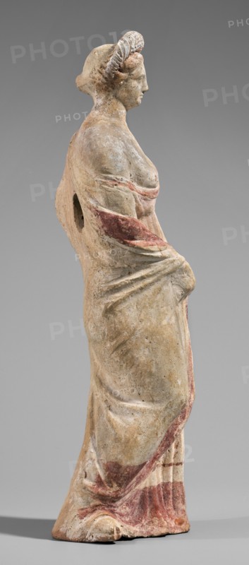Figurine of a maenad. A young woman standing in a wreath tied with a ribbon is dressed in a chiton slipped from her right shoulder exposing her right breast and upper arm, and a cloak that envelops her hands. The left hand rests on her hip; the right leg is pushed back. The robe is light pink decorated at the bottom with a wide border and a narrow beetroot-colored stripe above it. A similar border is found at the bottom, and a wide border in the same color runs around the cloak. The back of the figurine is unfinished. Without base. 3rd–2nd century BC. Myrina. Terracotta figurine. Height 26.5 cm, width 9.2 cm, depth 7.4 cm, weight 0.39 kg., A standing statue of a woman shown in profile facing right, rendered in a weathered pale beige material with traces of muted red pigment on the drapery and hair, wearing a long flowing garment with visible folds gathered at the waist; the figure has coiled hair or a braided headpiece, a defined nose and chin, and a rounded shoulder with an opening at...