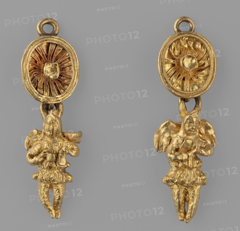 Two earring pendants in the shape of winged Geniuses, Pair of pendants - each composed of 2 parts: 1. Cups - suspended sideways, surrounded by profiled filigree and filled with a rosette with filigree, fastened with a high fitting for a stone missing; the bottom of the cup is reinforced with a soldered strip with a flatly soldered earring at the top and a perpendicularly soldered earring at the bottom. 2. Winged genius, - suspended by the lower earring handle, dressed in a short tunic chiton with hands joined at the front on the chest. The figurines are cast, with soldered wings cut from a single piece of sheet metal each, reinforced with crosswise soldered strips with an eyelet for hanging from a rosette. 1st half of the 3rd century BC, Greece; jewelry; weight 10 g., The image shows a pair of gold-colored dangling ornaments against a gray background, each with an oval top piece featuring a radial, textured central motif and a small loop at the top, connected by a short link to a small...