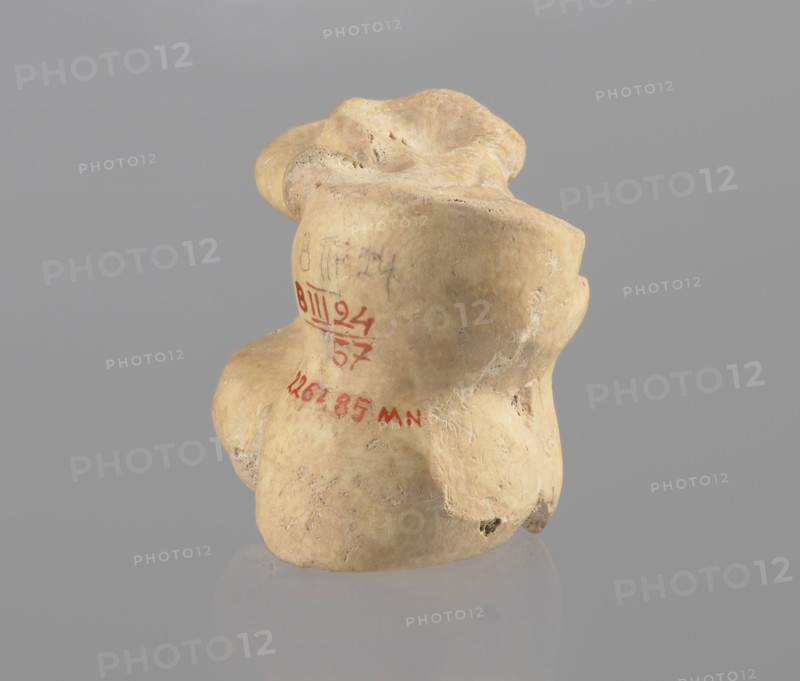 Astragalus of a cow, Myrmekion Crimea, Hellenistic period, astragalus condylo-talus bone of a cow, bones, antiquity, 323–30 BC, found at Myrmekion, product of nature, height 4.6 cm, A beige, irregularly shaped object with a textured, slightly pitted surface sits against a smooth gray background; the object has rounded lobes and depressions with subtle shadows, and its surface shows areas of lighter and darker beige along with small pale white marks and tiny dark spots.