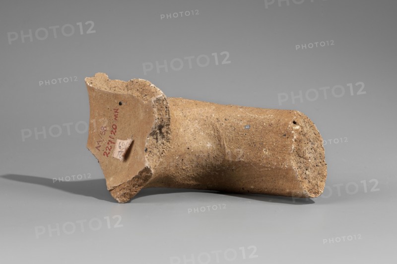 Fragment of the spout and vice of an amphora with a stamp, Papes c. 255–205 BC. Fragment of a Sinopean amphora: preserved spout, neck, and stamped vice. Spout is a semicircular roller; vice is oval in cross-section, flattened. Rectangular four-line stamp; to the right of the legend at height of lines 3–4 an emblem of a grapevine. First line contains the astynomic titulary; second line gives the astynomeus Eucharistos in generic form; third line his patronymic; fourth line the manufacturer name Papes in generic form. Attributed to M. Matera; 2nd half of 3rd century BC. Findspot: Myrmekion; origin: Sinope. Attributes: Sinopean stamp, vice; height 9.2 cm., A broken cylindrical clay fragment lying on its side on a smooth surface, with rough, uneven edges and a flared rim at one end; the fragment is predominantly brown and tan with speckles of darker brown and small lighter flecks, and it casts a soft shadow on the gray background.