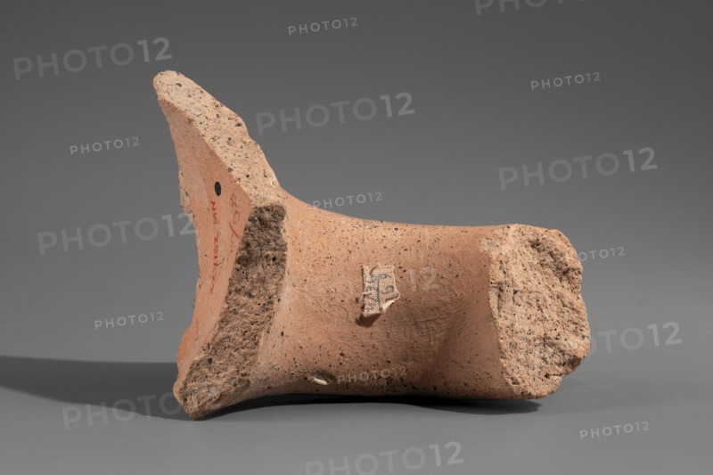 Fragment of a Sinopean amphora vice with a rectangular stamped impression inscription reversed, read right to left preserving the name of the Sinopean manufacturer Sagaris with patronymic; an emblem in the form of a leaf originally stood at the left of the stamp. Hellenistic period, ca. 320–300 BC. Found at Myrmekion Crimea. Stamp; height 8 cm. Made in Sinope. Maker attributed: Sagaris son of Numenius., A broken terracotta fragment rests on a flat surface against a neutral gray background, showing rough, porous edges and smoother inner surfaces. The piece is primarily a warm orange-brown color with darker speckles scattered across its surface and a few areas of lighter, beige-toned roughness where the ceramic is fractured. A small patch of fabric-like material is attached to the smoother face, displaying a mix of off-white and darker threads. Shadows beneath the fragment are muted gray, and there is a small round dark spot visible on one side of the piece.