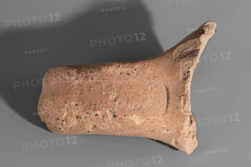 Fragment of a Sinopean amphora vice with a rectangular stamp impression on the upper surface. The fragmentarily preserved, reversed inscription read right to left contains the name of the Sinopean manufacturer, Sagaris, given with his patronymic; an emblem in the form of a leaf originally appeared at the left of the stamp. Dated ca. 320–300 BC. Origin: Sinope; findspot: Myrmekion. Height 8 cm. Maker: Sagaris son of Numenius., A fragment of a rough, earthen object with a textured, pitted surface in shades of tan and light brown rests against a smooth gray background; the object has an irregular, slightly cylindrical form with a flared, broken end and a rounded opposite end, showing a mix of darker speckles and small porous holes across its surface and casting a soft shadow on the background.