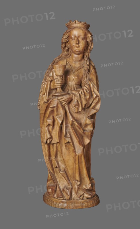 Figure of St. Barbara, sculptor unknown. Medieval wall sculpture, hollowed out from behind the head also has a circular hollow. Depicts St. Barbara in contrapposto, creating an S-shaped silhouette. Slightly oversized head on a cylindrical neck; elongated face with high forehead, straight narrow nose, full cheeks; small smiling mouth creating dimples in cheeks and a dimple in the chin. Long hair framed in thin parallel wavy strands, falling down her back with a strand over her right shoulder. Wears a crown finial broken. Right hand grasps the stem of a chalice attribute of St. Barbara; left hand, fingers covered by cloak, supports the chalice from beneath. Dress with triangular neckline and long wide sleeves, cinched at the waist with a decorative belt; voluminous cloak with numerous folds and dry thin ridges. Pointed left shoe peeks from beneath folds. Base round and irregularly carved. No polychrome. Circa 1500, possibly Silesia. Sculpture; height 102.0 cm, width 35.0 cm, depth 24.0 c...