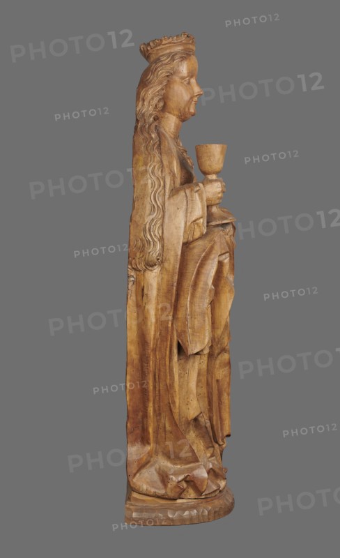 Figure of St. Barbara, medieval wall sculpture, hollowed out from behind head also has a circular hollow. Depicts St. Barbara in contrapposto, creating a slight S-shaped silhouette. Slightly oversized head on a cylindrical neck; elongated face with high forehead, straight narrow nose, full cheeks, small smiling mouth with dimples and a dimpled chin. Long hair framed and falling down her back, one strand draped over her right shoulder, carved in thin parallel wavy strands. She wears a crown finial broken. Right hand grasps the stem of a chalice; left hand, fingers covered by a cloak, supports the foot of the chalice. Dressed in a gown with triangular neckline and long wide sleeves, cinched with a decorative belt; voluminous cloak with numerous folds and dry thin ridges. Pointed left shoe peeks from beneath the folds. Round, irregularly carved base. No polychrome. ca. 1500, possibly Silesia. Sculpture, height 102.0 cm, width 35.0 cm, depth 24.0 cm., A wooden carved figure shown in profil...
