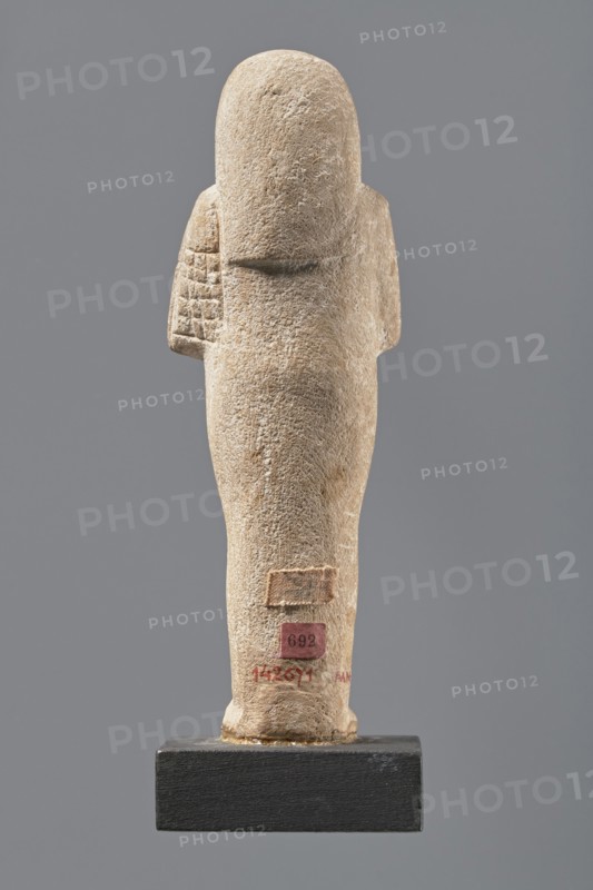 Ushabti, Mummy-shaped figurine made of limestone. Arms crossed on the chest, the right folded over the left, holding hoes in each hand. Rectangular basket on the back, slung by a single string over the left shoulder. Three-part smooth wig; almond-shaped eyes, wide nose, prominent mouth in a slight smile. Engraved inscription in a single column on the front of the legs; name illegible. Inscription partial: Shd Osiris mistress of the house of Naa, justified by her voice. ca. 1295–ca. 1186 BC; New Kingdom, 19th Dynasty, Egypt. Shabti. Height 13.3 cm, width 4.3 cm., A standing carved figure viewed from behind on a rectangular base, made of a light beige stone with a rough textured surface; the figure has a rounded head and a narrow torso with a few shallow incised rectangular patterns on one side suggesting a wrapped or detailed arm area, and subtle variations in the stone showing slightly darker beige and tan patches; the background is a flat mid-gray and the base is a dark charcoal color...