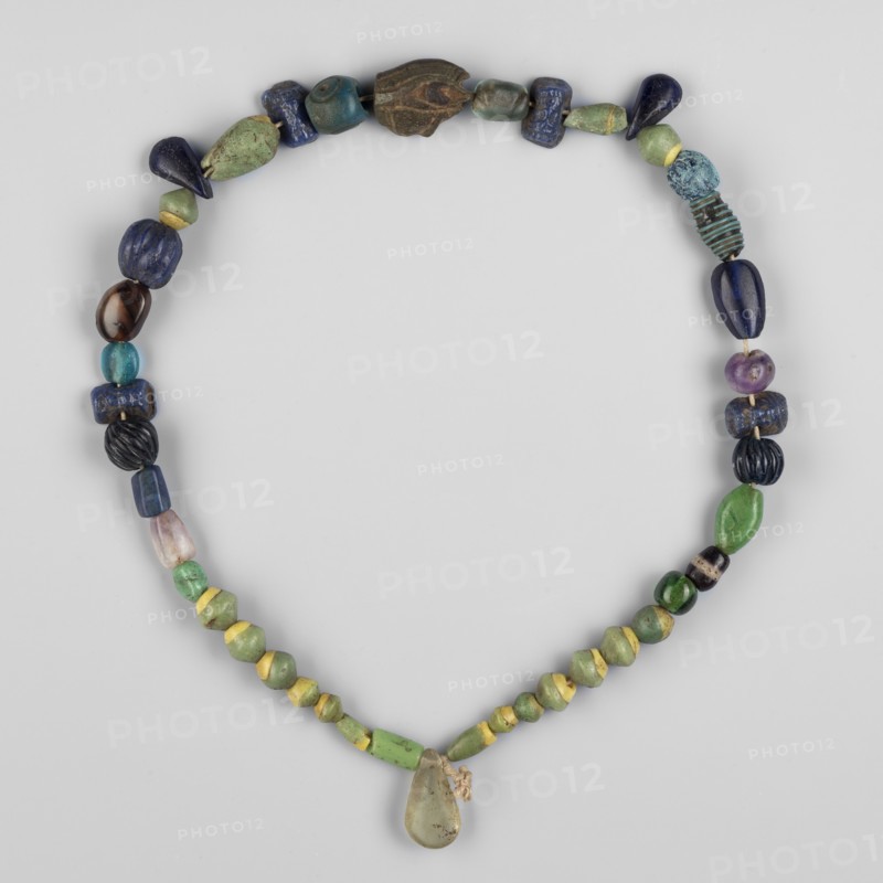 Necklace of 43 different beads, Goluchów Greater Poland Voivodeship. Scaraboids, udjat eyes, ornaments. Necklace consisting of 43 beads, including an udjat eye and 4 glass beads depicting a bearded face. Among the others, biconical and cylindrical beads made of green glass combined with yellow predominate, but many other shapes and colors can be found here - teardrops of black obsidian, an amethyst ball, melon-shaped beads made of dark glass, a quartz flower petal and many others. 664 BC – 395, Egypt; jewelry, amulet; length 38 cm., A necklace composed of a group of beads arranged in a loop, featuring a teardrop-shaped pendant at the bottom tied with a small cord; the beads show a variety of shapes including rounded, elongated, faceted, and ribbed forms. The visible colors include shades of green, blue, yellow, purple, black, brown, turquoise, and teal, with some beads showing mixed or mottled tones. Several beads have a smooth glossy surface, a few have matte or weathered textures, an...