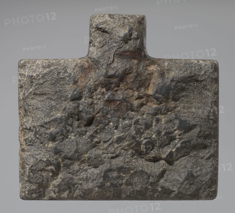 Nesichnum's sacrificial table; sacrificial formulas, vases, breads, cartouches, Nesichnum ca. 332–330 BC; ritual vessels iconographic; hieroglyphs; Ptolemaic Period, Egypt 332–30 BC; sacrificial table; height 5.5 cm, width 23 cm, depth 23 cm., A roughly rectangular stone slab with a small raised rectangular projection at the top, showing a coarse, uneven surface texture with pitted areas and angular indentations; the stone displays a mix of gray tones with darker charcoal patches and subtle brownish and rusty ochre areas, and the background behind the stone is a smooth neutral gray.