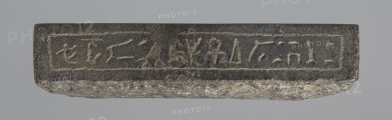 Nesichnum's sacrificial table; sacrificial formulas, vases, breads, cartouches, Nesichnum ca. 332–330 BC; ritual vessels iconographic; hieroglyphs; Ptolemaic Period, Egypt 332–30 BC; sacrificial table; height 5.5 cm, width 23 cm, depth 23 cm;, A rectangular stone slab with a rough, irregular lower edge rests against a neutral gray background; the top face is darker with a slightly textured surface and shows the impression of a framed rectangular panel across its length, while the lower exposed edge reveals lighter gray and beige mineral variations.