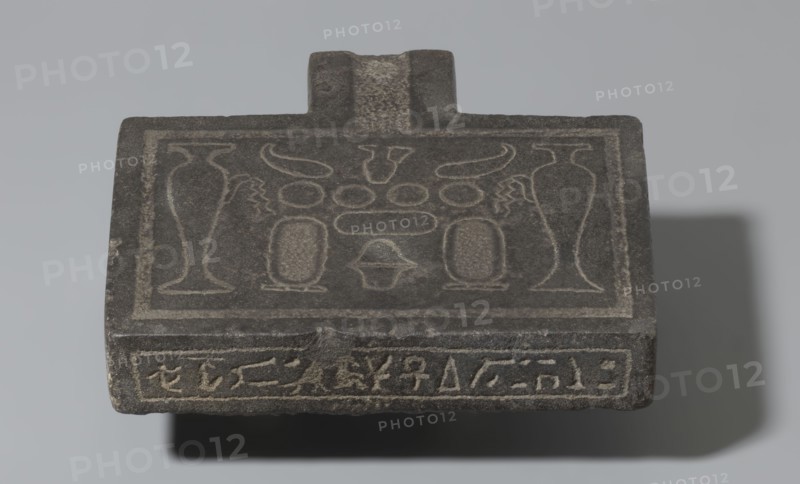 Nesichnum's sacrificial table; sacrificial formulas, vases, breads, cartouches, Nesichnum ca. 332–330 BC; ritual vessels iconographic; hieroglyphs; Ptolemaic Period, Egypt 332–30 BC; sacrificial table; height 5.5 cm, width 23 cm, depth 23 cm., A flat, rectangular stone object with a raised handle-like protrusion at one edge, showing carved linear designs and shallow relief motifs across its top surface; the stone appears predominantly dark gray with lighter gray incised lines highlighting the decorative patterns, and a shadow cast behind and below the object in darker gray tones.