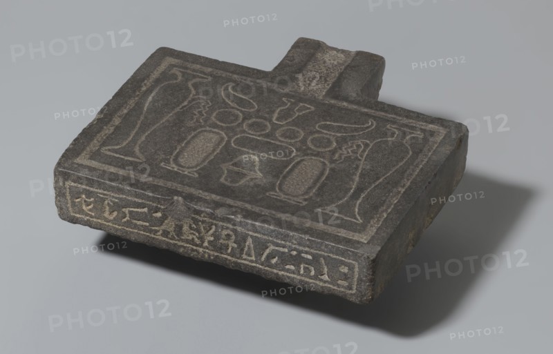 Nesichnum's sacrificial table; sacrificial formulas, vases, breads, cartouches, Nesichnum ca. 332–330 BC; ritual vessels iconographic; hieroglyphs; Ptolemaic Period, Egypt 332–30 BC; sacrificial table; height 5.5 cm, width 23 cm, depth 23 cm, A dark gray rectangular stone slab with a short protruding tab rests on a light gray surface, showing lighter carved outlines on its top face that form a symmetrical arrangement of abstract shapes and vessel-like forms; the slab's edges are worn and reveal a few rough brownish spots, and the object casts a soft shadow to the right.