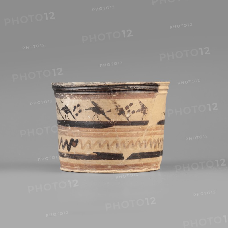 Pyxis with a duck motif. Greek, Boeotian, early 7th century BC. Clay yellowish/light gray to very pale brown with brown varnish and dark brown matte paint; red paint light reddish brown. Vessel with flat bottom, walls slightly flaring toward a profiled rim. Banded decoration: upper band shows a row of birds walking to the right in silhouette with lowered heads and raised wings; dotted rosettes between pairs of birds. Below, decoration enclosed in two wide dark bands consisting of two watered-firnis stripes and a zigzag line. Dimensions: depth 5.5 cm; diameter 7.3 cm; wall thickness 0.4 cm; height 6.5 cm; width 8.8 cm; weight 0.098 kg., A short cylindrical ceramic vessel sits against a plain gray background, showing a beige to light tan base color with decorative bands in darker brown and black. The rim is slightly flared and darker, with visible wear and chipping revealing lighter beige beneath. Around the upper part of the vessel are dark painted motifs including abstract birdlike sha...