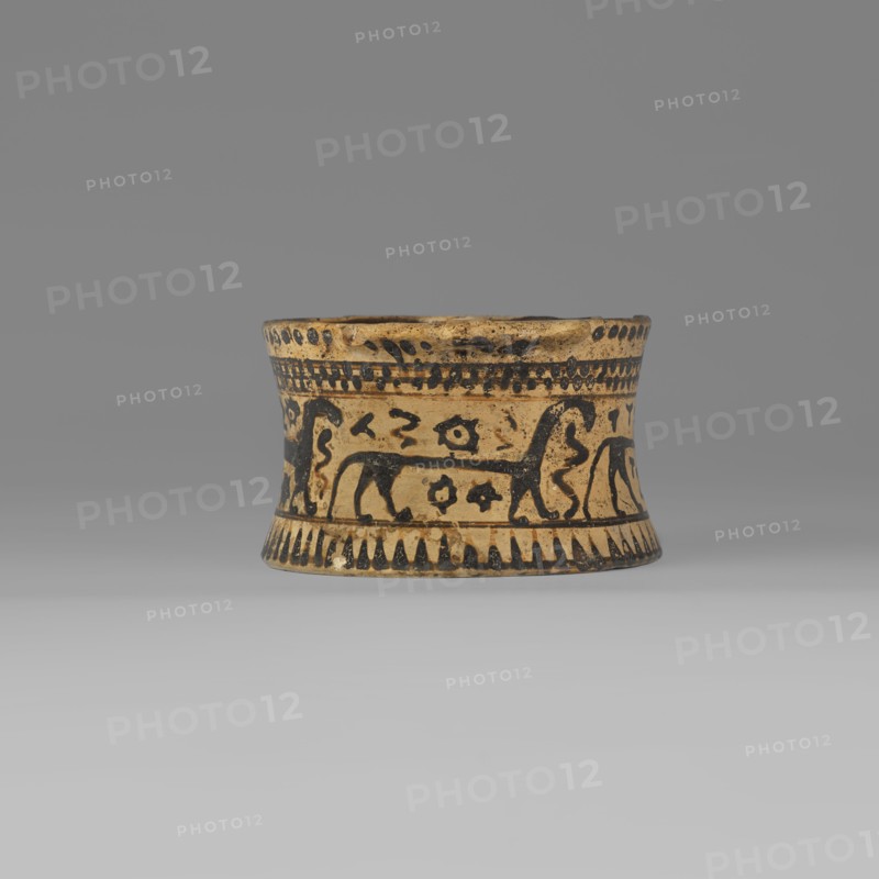 Orientalising pyxis with an animal frieze in the Silhouette style, images of animals, Middle Corinthian period 590–570 BC, cats. Thin-walled pyxis with a straight rim, slightly concave body walls, and a flat bottom. Small vices mounted horizontally just near the edge of the rim. The main decorative band, bounded at the top and bottom by a thin line of varnish, depicts figures of animals executed using the silhouette technique. Between the silhouettes there are simplified filling ornaments. Clay color: 5 Y 8/2 pale yellow Varnish color: 5 Y 2.5/1 black Corinth, pyxis vessel. Height 5.3 cm, width 10.2 cm, depth 8.5 cm, weight 0.102 kg., A short, flared cylindrical ceramic vessel sits centered against a neutral gray background; its body is a warm tan with dark brown or black painted decorations that form bands around the top and bottom edges and a central frieze. The painted frieze shows stylized animal figures and abstract motifs rendered in the dark color, repeating around the vessel, a...