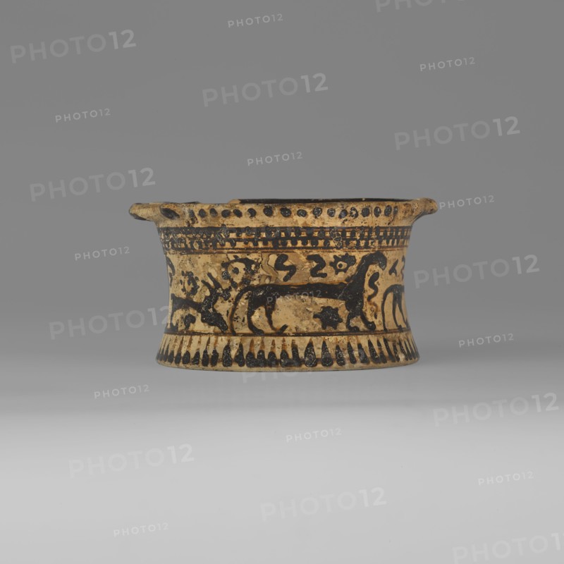 Orientalising pyxis with an animal frieze in the Silhouette style, images of animals, Middle Corinthian period, cats. Thin-walled pyxis with a straight rim, slightly concave body walls, and a flat bottom. Small vices mounted horizontally just near the edge of the rim. The main decorative band, bounded at the top and bottom by a thin line of varnish, depicts figures of animals executed using the silhouette technique. Between the silhouettes there are simplified filling ornaments. Clay color: 5 Y 8/2 pale yellow Varnish color: 5 Y 2.5/1 black 590–570 BC Middle Corinthian period, Corinth, pyxis vessel, height 5.3 cm, width 10.2 cm, depth 8.5 cm, weight 0.102 kg, A shallow, round ceramic vessel in beige and black sits centered against a neutral gray background, with a flared rim and a slightly concave body. The surface shows painted black decorative bands and motifs, including stylized animal silhouettes and abstract shapes encircling the midsection, a band of alternating dash-like marks a...
