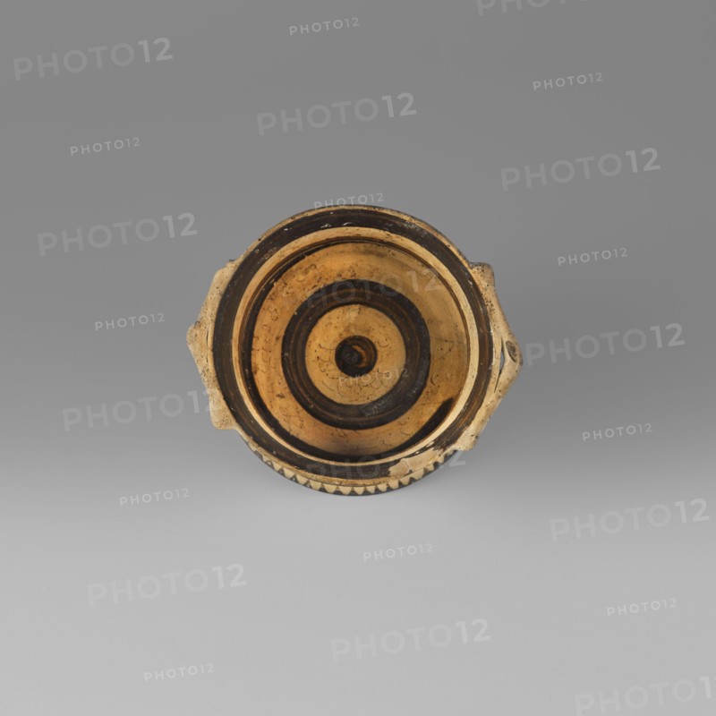 Orientalising pyxis with an animal frieze in the Silhouette style, Images of animals, Middle Corinthian period, cats. Thin-walled pyxis with a straight rim, slightly concave body walls, and a flat bottom. Small vices mounted horizontally just near the edge of the rim. The main decorative band, bounded at the top and bottom by a thin line of varnish, depicts figures of animals executed using the silhouette technique. Between the silhouettes there are simplified filling ornaments. Clay color: 5 Y 8/2 pale yellow Varnish color: 5 Y 2.5/1 black 590–570 BC dated to the Middle Corinthian period, Corinth. Pyxis vessel. Height 5.3 cm, width 10.2 cm, depth 8.5 cm, weight 0.102 kg., A round shallow vessel viewed from above with a circular interior decorated with concentric rings; the vessel is primarily tan and black with a warm yellowish-tan interior and dark black rings, a few worn areas showing mottled tan and brown, and two small opposed handles protruding from the rim; the exterior rim show...