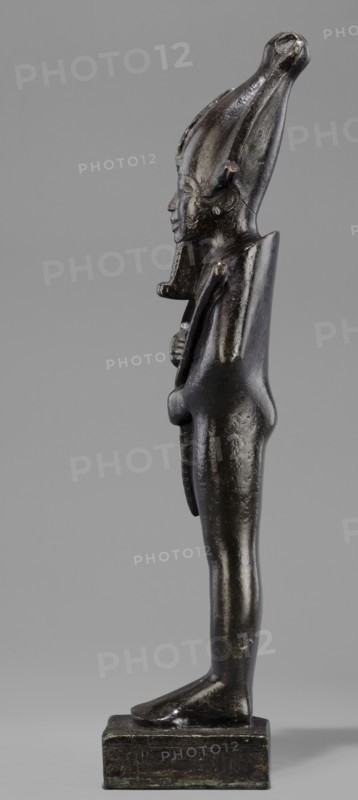 Figurine of the god Osiris. Statuette of a standing Osiris wrapped in a long, tight-fitting robe forming a characteristic rounding at the back of the shoulders. On the head is an atef crown with horns and a uraeus. The feet rest on a rectangular base, hollow inside, with a tenon for seating on the base. The four sides of the base are covered with a hieroglyphic inscription in two lines. Osiris mythology; Egyptian deities; mummy-shaped figurines; Egyptian art; Egyptian sculpture. 664–525 BC; Late Period, 26th Dynasty, Egypt. Statuette figurine. Height 31.5 cm; width 9.4 cm., A dark metallic figurine stands upright on a rectangular base against a plain gray background, showing a profile view of a humanlike figure with an elongated headdress that curves upward and backward. The figure's surface is glossy with reflections and areas of lighter sheen, and the overall color appears dark bronze with subtle greenish patina in places. The face has a pronounced nose and stylized features, and a s...