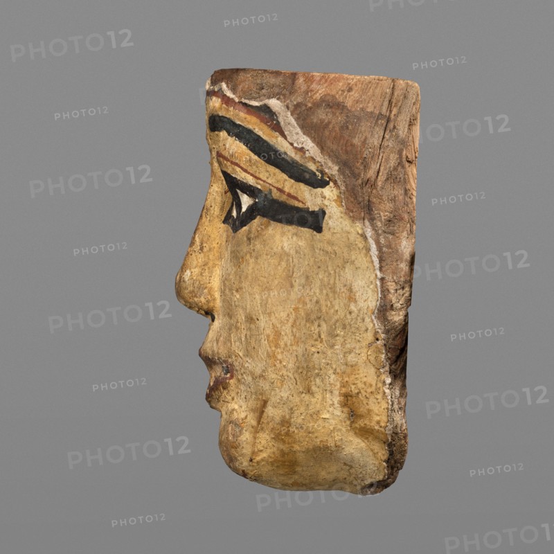 Woman's sarcophagus mask. Mask of wood covered with a layer of canvas with paint applied. The face is wide, painted yellow. Large almond-shaped eyes with pupils highlighted in black and arched eyebrows. Straight nose flattened at the tip. Mouth and chin protrusions marked in red, as is the outline of the eye sockets above the eyes. Face height: 15 cm. Sarcophagus fragments; masks, faces; grave goods. Mask from Egypt, 1st–2nd century; Roman period 4th century AD. Sarcophagus mask; height 15 cm, width 14.3 cm, depth 8 cm, weight 0.454 kg., A carved wooden profile of a human face oriented to the left with visible contours of a nose, lips, and chin, painted in a muted yellow-beige tone with areas of darker brown weathering; a painted eye area features bold black lines and a small white triangular highlight, with a band of reddish-brown and a thin yellow stripe above the eye; the top and back edges of the wood show natural brown wood grain and rough texture, and the background is a uniform ...