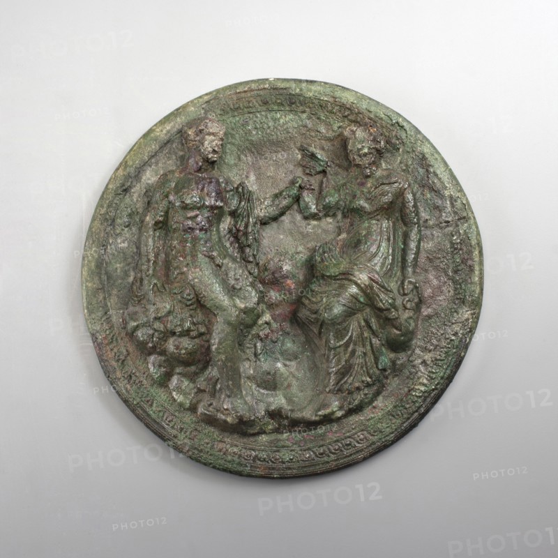 Mirror cover with an image of a couple. The bronze cover is decorated with a figurative scene of two people on the outside. The edge is also decorated with a braided ornament. The depiction includes a man and a woman facing each other, seated on rocks. The man's body is naked, with a chlamys draped over his left shoulder. The figure rests on a rock with his right hand, while his left touches the woman's right wrist. Dressed in a long chiton and himation, the woman also holds her left hand on a rock. The robe forms a few wide folds around the hips. A series of thick folds of fabric are also visible on the legs. Higher up, the fabric accentuates the woman's natural shape, while the folds are sparse and subtle. The depiction can be interpreted as a pair of lovers. Circular holes are carved in the man's right foot and the woman's left foot. Circular, c. 300 BC, Corinth; mirror; height 3 cm, diameter 14 cm., A round, relief-carved disc with a weathered, rough surface showing shades of green...