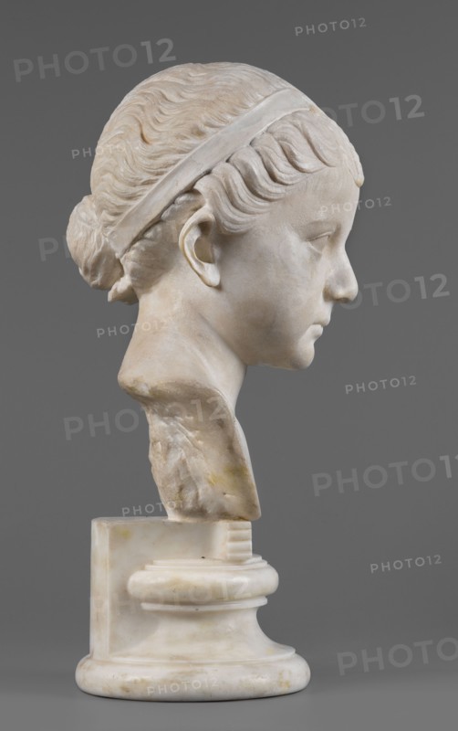 Portrait of a princess from the Julio-Claudian dynasty Julia?, unknown Roman workshop. The bust depicts a girl with a round, rather chubby face, turned slightly to the right; a rounded, elongated chin, and small, slightly pursed lips with a pronounced, wavy line of the upper lip. The almond-shaped, smooth eyes are rather shallowly set, with marked upper eyelids and barely visible eyebrows. The ears are carefully styled. The wavy hair is combed up and tied with a headband. At the back of the head, it is tied in a bun and a braid that falls down the middle of the head onto the smooth forehead. End of the 1st century BC–beginning of the 1st century AD, Roman Empire 27 BC–476. Sculpture. Height 25.7 cm, width 16 cm, depth 12.5 cm, weight 11.5 kg., The image shows a marble bust of a young person in profile against a solid gray background; the sculpture displays a smooth pale off-white surface with subtle beige and yellowish discolorations, carved wavy hair pulled back into a bun and held by...