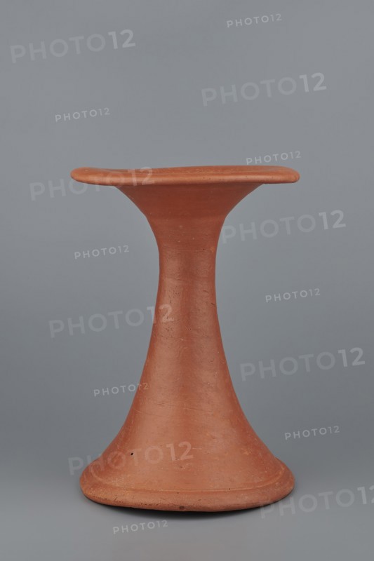 Bowl base. Base for a vessel with an almost equal diameter of both rims; both rims are irregular, with a wavy line. The center of the base is empty. The narrowest part of the base is approximately 2/3 of the height of the entire vessel. Made of Nile clay NB1. Surface leveled, covered with an engobe, then polished. Vessel color: red. Complete, very well preserved. c. 2494–c. 2181 BC; Old Kingdom, 5th–6th Dynasty, Edfu. Vessel; top diameter 17.5 cm, bottom diameter 18 cm, height 25.6 cm, weight 1.30 kg., A terracotta object shaped like a tall hourglass with a flared base and a flat, slightly rounded top is centered against a smooth gray background; the object has an earthy reddish-brown color with a matte, slightly rough surface showing small imperfections and subtle variations in tone, and the background displays a uniform cool gray.