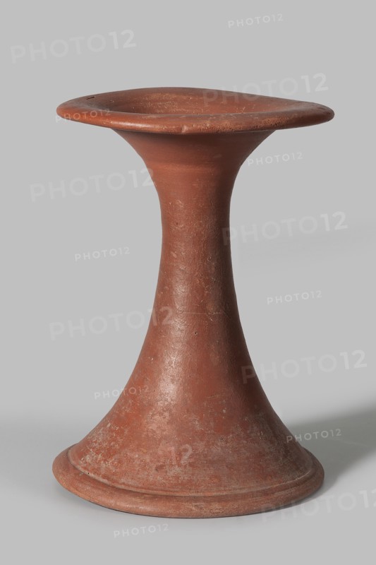 Bowl base, Base for a vessel with an almost equal diameter of both rims; both rims are irregular with a wavy line. The center of the base is empty. The narrowest part of the base is approximately 2/3 of the height of the entire vessel. Vessel made of Nile clay NB1. The surface is leveled, covered with an engobe, and then polished. Vessel color: red. Complete vessel, very well preserved. c. 2494–c. 2181 BC; Old Kingdom, 5th–6th Dynasty, Edfu. Vessel; top diameter 17.5 cm, bottom diameter 18 cm, height 25.6 cm, weight 1.30 kg., A single reddish-brown terracotta object with a flared circular base tapering into a narrow waist and then widening into a shallow, rounded top rim, showing a worn, slightly rough surface texture with areas of lighter discoloration and small surface imperfections, set against a plain light gray background.
