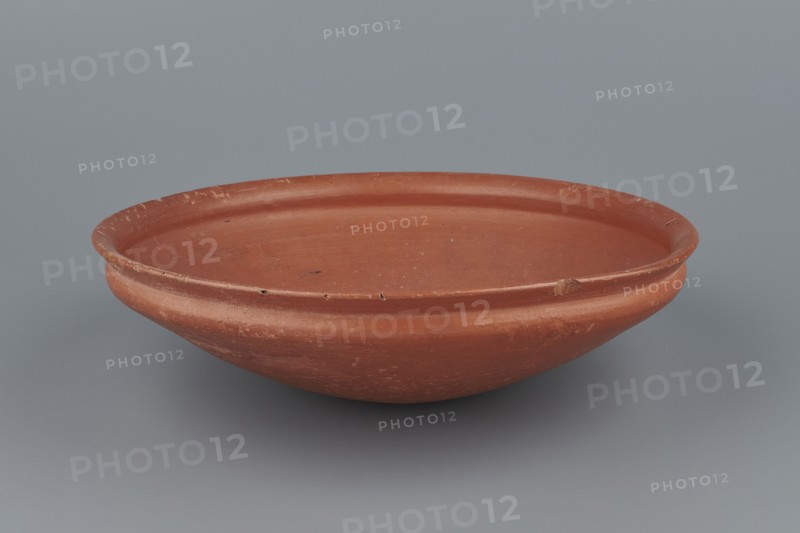 Medum-type bowl, Egyptian pottery, Medum-type bowls. Clay vessel with hemispherical shape narrowing just below the rim to form a carination; rounded bottom; straight, slightly widening spout whose diameter is slightly larger than the shoulders. Surface even, covered with red engobe and polished. Vessel color: red. Fairly well preserved with minor losses on the rim. c. 2494–c. 2181 BC; Old Kingdom, 5th–6th Dynasty, Edfu. Vessel; height 7 cm; diameter 26.4 cm spout; weight 0.90 kg., A shallow round dish with a raised rim in terracotta red, showing a smooth matte surface with areas of lighter wear and small dark spots, set against a cool gray background.