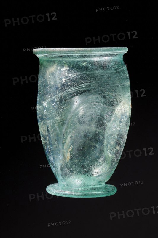 Glass cup;, Egypt, 1st–2nd century AD. Type: Isings form 32. Transparent bluish-green glass. Cup with a slightly flaring spout, decorated with four oval concavities on the belly. Ring-shaped foot with a clearance created by pressing the bottom inward; free-blown. Outer diameter of spout: 6.10 cm. Height: 9 cm. Belly diameter: 6.70 cm. Outer diameter of bottom: 4.60 cm. Vessel; weight 0.054 kg., A translucent glass vessel with a rounded body and a slightly flared rim sits against a black background, showing a bluish-green color with areas of clearer and cloudy glass, surface scratches, and a few small whitish encrustations; the base is a flattened circular foot of the same bluish-green glass and the overall surface displays swirling internal striations and variations in opacity.