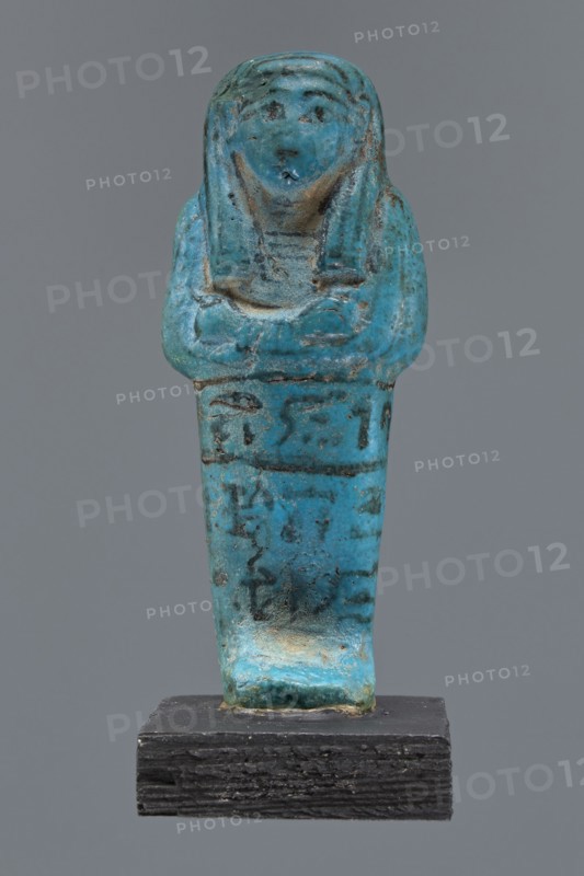 Ushabti Cha-em-uase. A mummy-shaped figurine shabti made of faience and pressed into a mold. Details of the face, jewelry, and bag are painted. Hands folded across the chest—right over left. Wide bracelets on the wrists. An ush necklace on the chest. No tools in the hands; a bag on the back. A three-part wig, combed into strips, with borders at the ends. Painted inscription, three lines of text on the legs: Shd Osiris Cha-em-Wese justified with his voice said: O — the beginning of the sixth chapter of the Book of the Dead. Ca. 1295–1069 BC; New Kingdom, 19th–20th Dynasty. Height 10.1 cm, width 4.1 cm. Maker, private name Cha-em-uase., A small statuette shaped like a standing human figure with folded arms, rendered in a bright turquoise-blue material with areas of darker blue and greenish patina and some brownish and blackened spots, mounted on a dark textured rectangular base; the figure has a rounded head with stylized facial features and a headdress-like form extending down the sides...