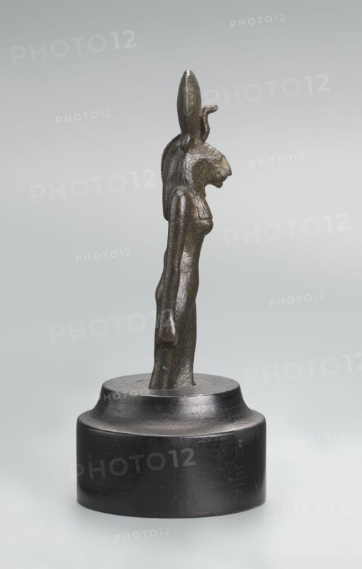 Sekhmet figurine, lion-headed goddess shown standing or walking with a lioness head in a tripartite wig topped by a solar disk with a uraeus. Right arm hangs at her side, clenched into a fist revealing a hole from a lost cult object; left arm broken at the elbow. Figurine broken at the knuckles and mounted on a modern cylindrical wooden base with contoured edge. 664–525 BC, Late Period, 26th Dynasty, Egypt; terracotta? material not specified; height 9.7 cm., A small metallic statuette depicting a standing anthropomorphic figure with an animal-like head and elongated ear-like elements on top, mounted on a round dark pedestal; the figure has a slender elongated torso, visible arms at its sides, and textured surface with worn highlights. The visible colors are dark bronze with greenish-brown patina on the figure and deep black on the pedestal, set against a pale gray to white gradient background.