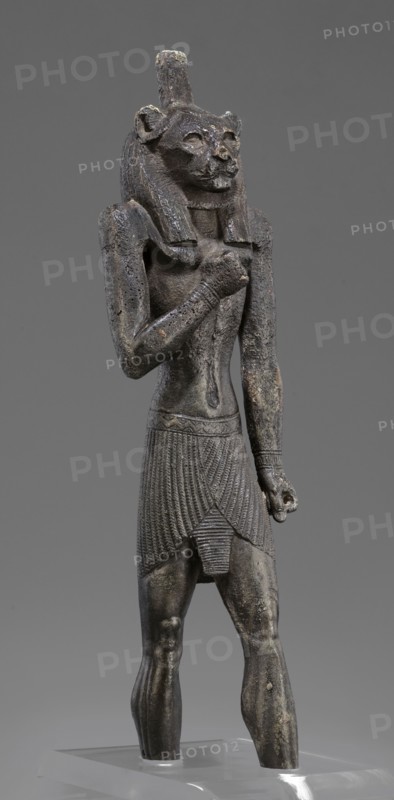 Figurine of the god Mahes. Figurine of a striding man with a lion's head. The lion's head is set directly on the shoulders. The wig is striped. A square pin is on top of the head. The right arm is bent at the elbow and placed on the chest, the left arm is freely placed along the body. A short striped loincloth is on the hips., Mahes myth.. Egyptian deities; Egyptian art; Egyptian sculpture. Late Period, 26th Dynasty, 664–525 BC, Egypt. Figurine; height 26 cm, width 7 cm, depth 3.8 cm., A dark gray statuette of a standing anthropomorphic figure with a feline head and a tall cylindrical crown, showing a textured mane and carved facial features, wearing a pleated skirt with detailed vertical and diagonal patterns and a narrow decorative belt, with one arm bent across the chest and the other hanging down holding a small rounded object, the surface showing areas of lighter wear and small patches of brownish discoloration, set against a uniform medium gray background and mounted on a clear b...