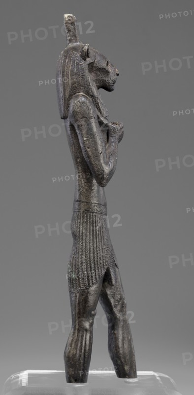 Figurine of the god Mahes, Mahes myth., Egyptian deities, Egyptian art, Egyptian sculpture. Figurine of a striding man with a lion's head. The lion's head is set directly on the shoulders. The wig is striped. A square pin is on top of the head. The right arm is bent at the elbow and placed on the chest, the left arm is freely placed along the body. A short striped loincloth is on the hips. 664–525 BC; Late Period, 26th Dynasty, Egypt. Figurine; height 26 cm, width 7 cm, depth 3.8 cm., A dark gray metal statue of a standing anthropomorphic figure with a feline head, wearing a long textured headdress and a pleated skirt, shown in profile against a neutral gray background; the figure's arms are held bent at the chest with hands clasped, the surface shows rough patina with hints of lighter gray and subtle greenish corrosion, and it stands on a transparent base with reflections.