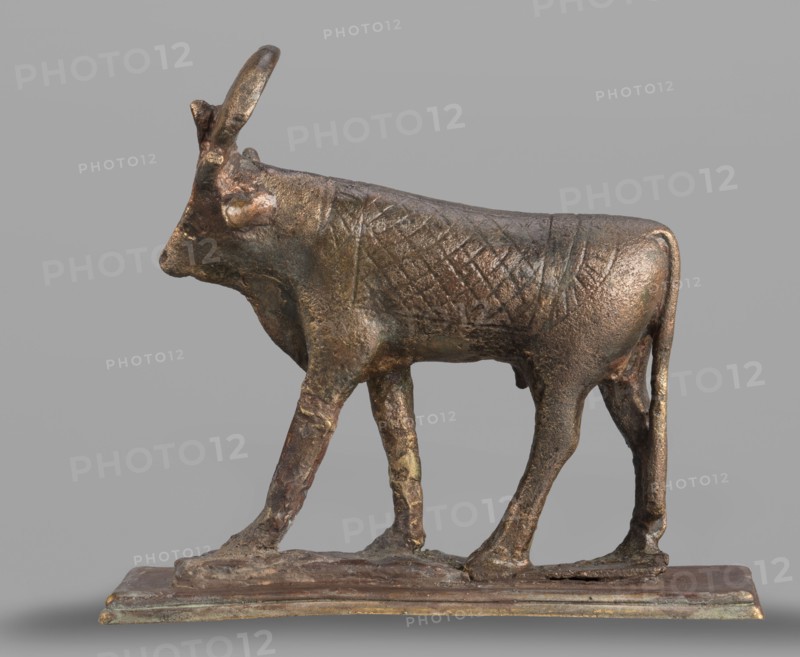 Apis bull figurine, sacred animal of the god Ptah, Apis mythology, bulls, sacred animals, animal figurines, Egyptian art, 664–525 BC; Late Period, 26th Dynasty, Egypt, figurine; height 7 cm, width 6 cm, weight 0.164 kg, A small bronze sculpture of a walking bull shown in profile on a rectangular base, with a textured body surface and visible wear; the bull has curved horns, a pronounced muzzle, and four slender legs positioned as if in mid-stride, and the colors visible include metallic brown, dark bronze, greenish patina, and some lighter goldish highlights against a neutral gray background.