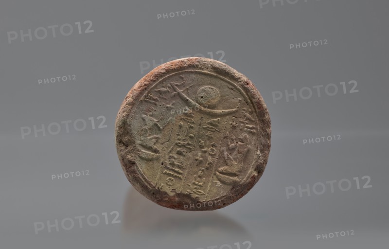 Basa's funeral cone, created by Basa ca. 664–610 BC. The hieroglyphic inscription on the base runs in two vertical columns. On either side a figure facing the inscription, kneeling on the neb sign, with hands raised in prayer. Above the inscription is a representation of a solar barque. Boat and solar disc iconography; hieroglyphs. Scen.Gal.Eg. Davies-Macadam, Corpus No. 602. Date: 664–610 BC; Late Period, 26th Dynasty reign of Psammetik I. Provenance: Western Thebes tomb 389 on Asasif. Cone-shaped funerary cone; height 9 cm, width 23 cm, weight 1.072 kg., A round, weathered object with a rough, uneven rim sits against a gray background; the object shows textured surfaces and carved or impressed shapes and markings across its face, with a raised circular border and a crescent-like curved line near the top, and the surface displays a mix of earthy tones including brown, reddish-brown, beige, and muted green.