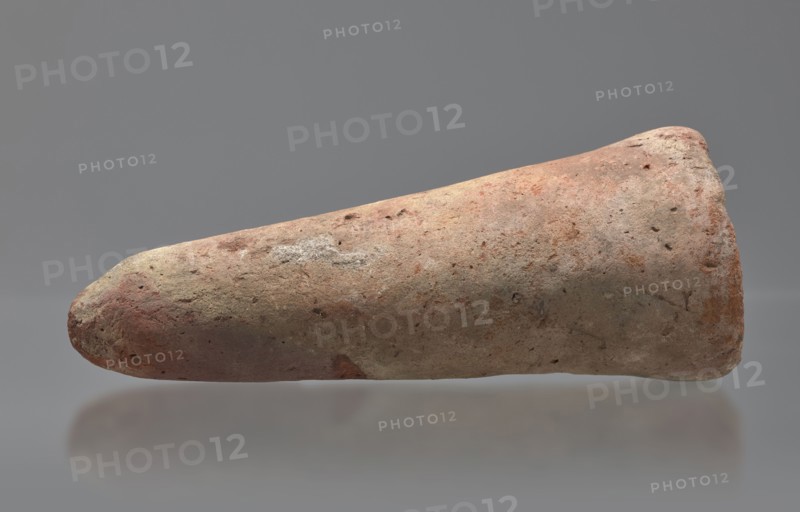 Basa's funeral cone, creator, Basa ca. 664–610 BC. Cone-shaped funerary cone, height 9 cm, width 23 cm, weight 1.072 kg. Late Period, 26th Dynasty reign of Psammetik I, Western Thebes tomb 389 on Asasif. The hieroglyphic inscription on the base runs in two vertical columns. On either side a figure faces the inscription, kneeling on the neb sign with hands raised in prayer. Above the inscription is a representation of a solar barque. Iconography: boat, solar disc; contains hieroglyphs. Davies-Macadam, Corpus No. 602., A single elongated, conical object rests horizontally against a smooth gray background with a soft reflection beneath it; the object has a rough, pitted surface showing a mix of earthy colors including tan, beige, reddish-brown, pinkish hues, and patches of light gray, with some darker spots and mottled texture across its surface.