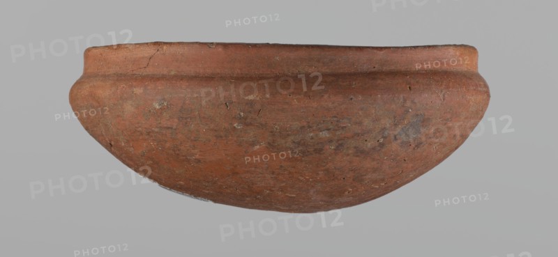 Medum-type bowl, small Medum-type bowl with characteristic carination under the rim. Rounded bottom. Arched walls, gently breaking towards the center of the vessel around 2/3 of its height. Straight, perpendicular rim. Narrow, sloping shoulders. The diameter of the rim is significantly smaller than the diameter of the vessel's arms. Clay probably NB2. Surface leveled and covered with a layer of engobe, then polished. Vessel color: red. c. 2686–c. 2181 BC; Old Kingdom, Edfu. Vessel; height 4.5 cm, diameter 11 cm rim, diameter 11.6 cm arms., A rounded, shallow ceramic bowl occupies the center of the image against a plain light gray background; the bowl is primarily reddish-brown with patches of darker brown and muted orange, and it shows a weathered, slightly rough surface texture with small pits and irregular marks, a rounded lip with a subtle ridge near the rim, and some areas of lighter beige and grayish discoloration scattered across the exterior.
