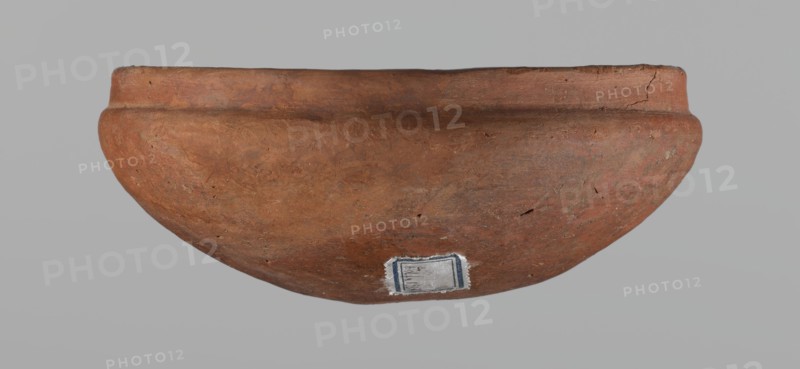 Medum-type bowl, A small Medum-type bowl with characteristic carination under the rim. Rounded bottom. Arched walls, gently breaking towards the center of the vessel around 2/3 of its height. Straight, perpendicular rim. Narrow, sloping shoulders. The diameter of the rim is significantly smaller than the diameter of the vessel's arms. Clay probably NB2. Surface leveled and covered with a layer of engobe, then polished. Vessel color: red. c. 2686–c. 2181 BC; Old Kingdom, Edfu. Vessel; height 4.5 cm, diameter 11 cm rim, diameter 11.6 cm arms., A reddish-brown rounded clay vessel with a slightly flared rim, showing surface texture, small chips, and cracks, set against a plain light gray background; a small rectangular patch with worn edges and a bit of dark framing is visible on the lower outer surface.