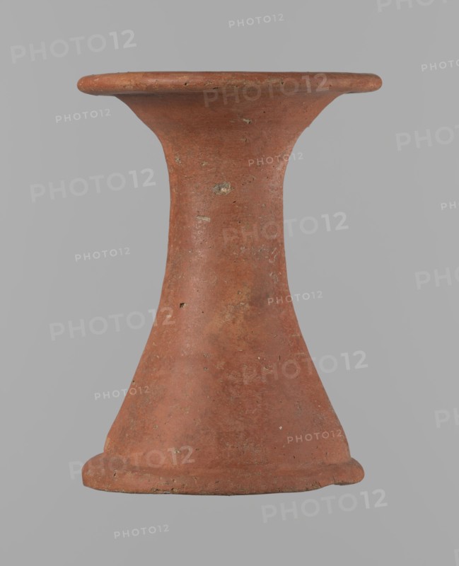 Bowl stand, Base for a vessel with an almost equal diameter of both rims. Both rims are irregular, with a slightly wavy line. The center of the base is empty. The narrowest part of the base is approximately 2/3 of the height of the entire vessel. Made of Nile clay NB2. The surface is leveled and covered with an engobe, then polished. Red in color. Complete, very well preserved. Set with bowl 138673. ca. 2494–ca. 2181 BC; Old Kingdom, 5th–6th Dynasty, Edfu. Vessel; height 13.7 cm; diameter 10.2 cm rim; diameter 9.3 cm bottom., An upright hourglass-shaped clay object with a flared rim and base, showing a weathered surface with small chips and rough patches, in shades of reddish-brown and terracotta against a flat gray background.
