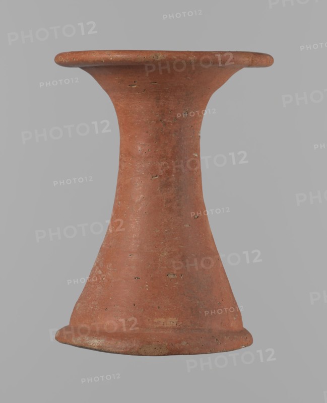 Bowl stand, Base for a vessel with an almost equal diameter of both rims. Both rims are irregular, with a slightly wavy line. The center of the base is empty. The narrowest part of the base is approximately 2/3 of the height of the entire vessel. Vessel made of Nile clay NB2. The surface is leveled and covered with an engobe, then polished. Vessel color: red. Complete vessel, very well preserved. Set with bowl 138673. ca. 2494–ca. 2181 BC; Old Kingdom, 5th–6th Dynasty, Edfu. Vessel, height 13.7 cm; diameter 10.2 cm rim; diameter 9.3 cm bottom., A single terracotta object shaped like an hourglass or spool, with a flared rim at the top and a flared base at the bottom, shown against a plain light gray background. The object is primarily reddish-brown with small patches of lighter beige and darker brown where the surface shows wear or texture. Its surface has a rough, slightly pitted texture with scattered small irregularities and tiny chips. The silhouette tapers inward at the midsection ...