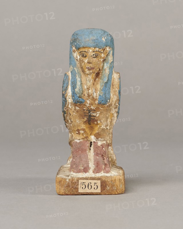 Figurine of the Soul of Ba, Ba mythology; birds, djed pillars, wigs, beards, feathers; The figurine has carefully carved and painted facial details and a reproduced arrangement of feathers on the wings. On the head is a tripartite wig painted blue. On the chest below the wig is a depiction of a djed pillar. Straps supporting a false beard are painted along the cheeks. The figurine's disproportionately large feet rest on a rectangular base. 664–332 BC, Late Period, Egypt; wood figurine, height 11 cm, width 9.5 cm, depth 4.5 cm., A small painted figurine of a seated human-like figure shown from the front, with a blue headpiece and blue accents along the sides of the head, a face painted in beige tones with darker details for features, a torso in worn beige and brown paint with traces of darker markings, and lower legs painted in muted reddish-brown sitting on a rectangular wooden base that is light brown; the background is a uniform light gray.
