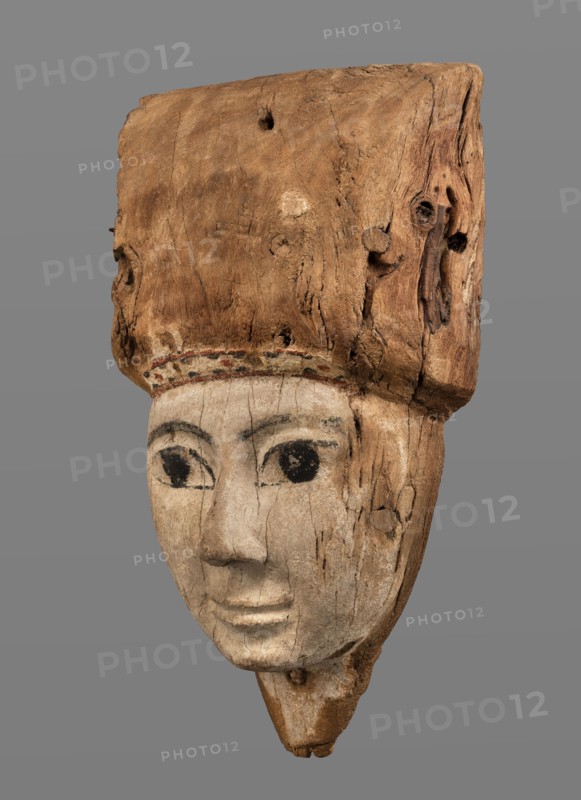 Sarcophagus mask, Sarcophagus mask with fragment of a high wig above the forehead, decorated on the edge of the forehead with a chain motif with details painted in black and red. The face is painted light cream; the large eyes and eyebrows are highlighted with black paint. Wig iconographic, sarcophagus fragments; masks, faces; grave goods. Face height: 12.5 cm. Overall height 31 cm, width 17 cm, depth 9 cm. Date: 332–30 BC, Ptolemaic Period, Egypt., A carved wooden head with a worn, light brown upper section and a pale, weathered lower face; the face shows painted dark eyes and dark eyebrow shapes, with visible cracks and grain lines across the wood; a narrow band of faint, muted color sits between the upper and lower sections; the upper wooden portion is rough, textured, and darker brown with knots and holes; the background is a uniform gray.