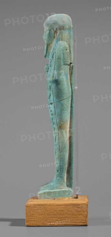 Udja-hera shabti, Udja-hera ca. 380–343 BC, wigs, hieroglyphs, hoes, beards facial hair, Book of the Dead, funerary figurine, grave goods, mummy-shaped figurine, standing on a base, back supported by a pillar. Arms crossed on the chest, right folded over left, elbows and shoulders subtly marked. Hoes in each hand; handles slightly curled. In the right hand is a twisted rope with a basket slung over the left shoulder. Convex tools, engraved basket. Long, three-part wig combed into narrow strands with main strands ending in borders. Long beard, slightly tapering and curled. Large ears. Proportionate, carefully crafted facial details; mouth set in a slight smile. Inscription engraved; arranged in three columns separated by lines on the front of the figurine's legs. 380–343 BC; Late Period, 30th Dynasty, Egypt. Shabti; height 15.5 cm, width 3.9 cm., A small turquoise-green figurine with a matte, slightly weathered surface stands upright on a rectangular wooden base; the figure is shown in ...