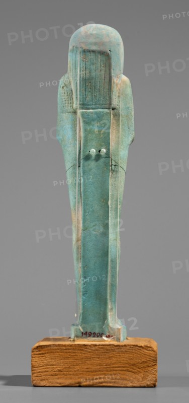 Udja-hera shabti, Udja-hera ca. 380–343 BC. Mummy-shaped funerary figurine shabti, Late Period, 30th Dynasty, Egypt. Standing on a base with back supported by a pillar; arms crossed on the chest right over left with elbows and shoulders subtly marked. Holds hoes in each hand with slightly curled handles; right hand holds a twisted rope from which a basket is slung over the left shoulder. Convex tools and engraved basket. Long three-part wig combed into narrow strands ending in borders; long slightly tapering curled beard; large ears; proportionate, carefully crafted facial details with a slight smile. Inscription engraved and arranged in three columns separated by lines on the front of the figurine's legs. Height 15.5 cm; width 3.9 cm., A weathered turquoise-green figurine stands upright on a rectangular wooden base, showing a stylized human form with a rounded head and elongated body; the surface has areas of lighter green and pale beige discoloration, small pits and abrasions, and su...