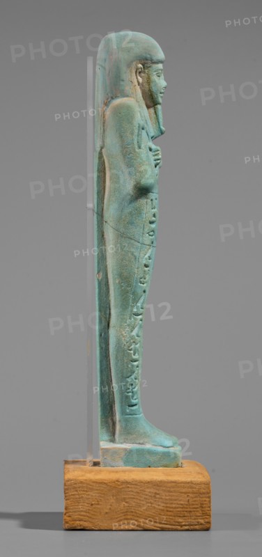 Udja-hera shabti, Udja-hera ca. 380–343 BC. Mummy-shaped funerary figurine shabti, Late Period, 30th Dynasty, Egypt. Height 15.5 cm, width 3.9 cm. Standing on a base with back supported by a pillar; arms crossed on the chest right over left with elbows and shoulders subtly marked. Holds hoes in each hand with slightly curled handles; right hand holds a twisted rope from which a basket is slung over the left shoulder. Convex tools and an engraved basket depicted. Wears a long three-part wig combed into narrow strands ending in borders; long slightly tapering, curled beard; large ears; proportionate, carefully crafted facial features and a slight smile. Inscription engraved in three columns separated by lines on the front of the legs. Materials/attributes: wigs, hieroglyphs, hoes, beards; function as grave goods linked to the Book of the Dead. Provenance: purchase., A small standing figure sculpture shown in profile on a wooden base, the figure is a greenish-blue with weathered lighter a...
