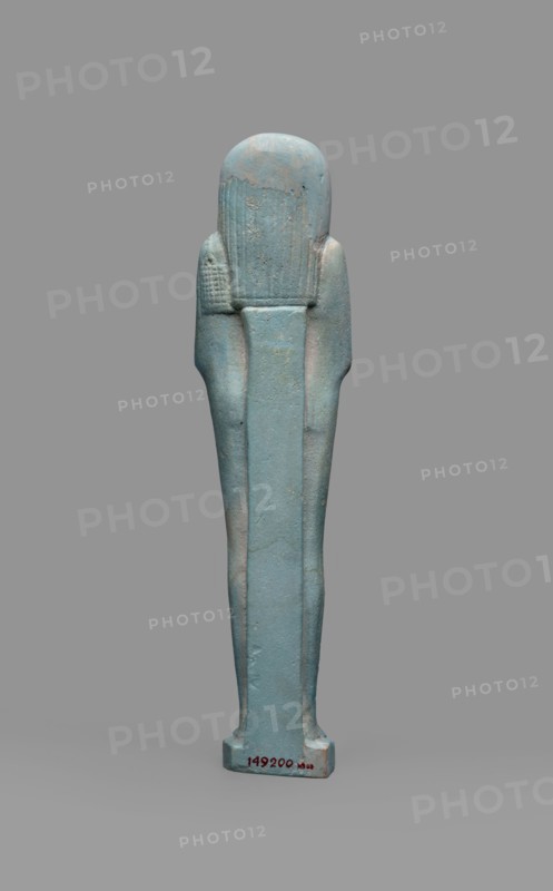 Udja-hera shabti, Udja-hera ca. 380–343 BC. Mummy-shaped funerary figurine shabti, Late Period, 30th Dynasty, Egypt. Height 15.5 cm, width 3.9 cm. Standing on a base with back supported by a pillar. Arms crossed on the chest, right folded over left; elbows and shoulders subtly marked. Holds hoes in each hand with slightly curled handles; right hand also holds a twisted rope from which a basket is slung over the left shoulder. Convex tools and an engraved basket. Long three-part wig combed into narrow strands ending in borders; long slightly tapering curled beard; large ears; proportionate, carefully crafted facial details with a slight smile. Inscription engraved in three columns separated by lines on the front of the legs. Mentions wigs, hieroglyphs, hoes, beards, Book of the Dead, funerary figurines, grave goods., A tall, vertical statuette with a smooth, elongated form in shades of blue and turquoise, showing areas of pale greenish-blue and subtle beige wear; the figure has a rounde...