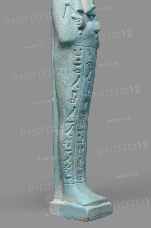 Udja-hera shabti, Udja-hera ca. 380–343 BC. Mummy-shaped funerary figurine shabti, Late Period, 30th Dynasty, Egypt. Height 15.5 cm; width 3.9 cm. Standing on a base, back supported by a pillar. Arms crossed on the chest, right folded over left; elbows and shoulders subtly modeled. Holds hoes in each hand with slightly curled handles. Right hand holds a twisted rope from which a basket is slung over the left shoulder; convex tools and engraved basket. Long three-part wig combed into narrow strands with main strands ending in borders. Long, slightly tapering curled beard; large ears; proportionate, carefully crafted facial details with a slight smile. Inscription engraved in three columns separated by lines on the front of the figurine's legs. Mentions wigs, hieroglyphs, hoes, beards, Book of the Dead, funerary figurines, grave goods., A turquoise-blue elongated figurine fragment showing the lower half of a standing human form with a few carved vertical grooves and a column of raised ca...