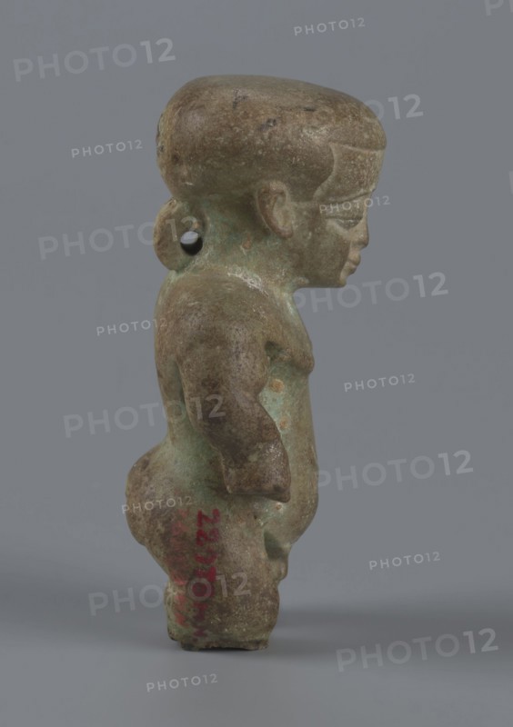 Amulet — Ptah-dwarf Pataikos, Figurine of a naked dwarf man with a large head with a wide forehead, short arms with clenched fists pressed to the hips, and short legs broken off at the knees. Carefully modelled. Ptah-dwarf mythology. 7th–4th century BC, Late Period, Egypt. Figurine/amulet. Height 6.5 cm; width 3.7 cm; depth 2.6 cm., A small figurine viewed in profile with a rounded head and a pronounced forehead, showing a facial profile with a closed mouth and a softly defined nose; the body is compact with rounded limbs and a slightly protruding belly, and there is a loop-like projection at the back of the head with a hole through it. The surface shows a mottled patina in shades of green and brown with areas of darker brown and some lighter greenish spots, and a cool gray background behind the figure.